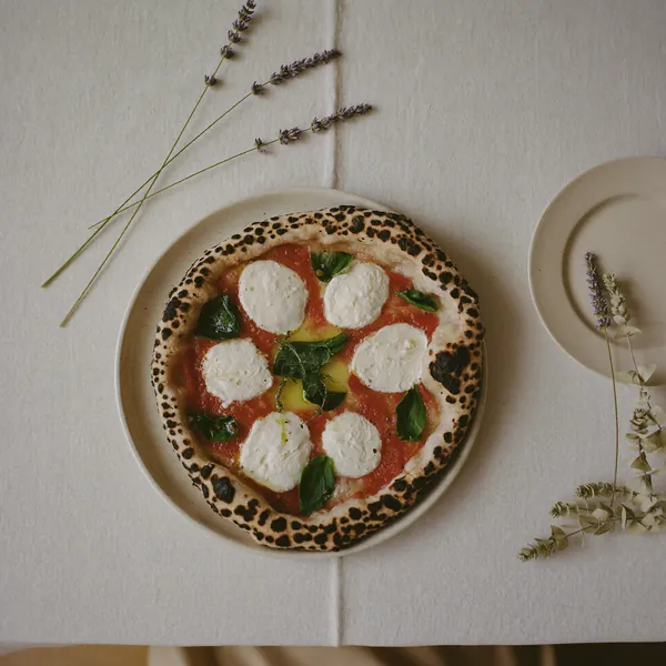 Een zuurdesempizza met mozzarella en basilicum, van bovenaf op crème linnen met lavendel ernaast.